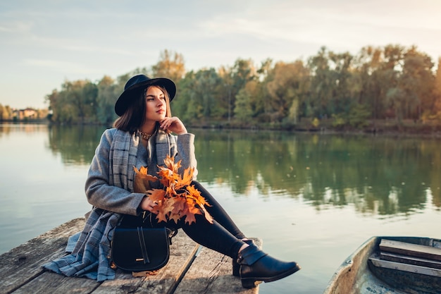 Frau, die sich am Seepier mit dem Boot entspannt und die Herbstlandschaft bei Sonnenuntergang mit Zweigen mit Blättern bewundert. Aktivitäten in der Herbstsaison