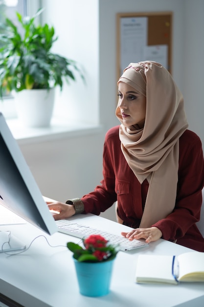 Frau, die sich beschäftigt fühlt. Dunkeläugige Frau mit Kopftuch, die sich beschäftigt fühlt, während sie in der Nähe des Computers sitzt