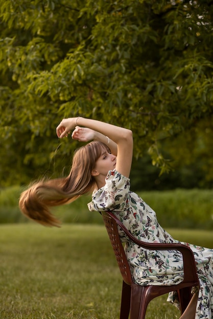 Foto frau, die sich im park entspannt, sitzt auf einem stuhl im wald mit langen blonden haaren. zartes ästhetisches mädchen