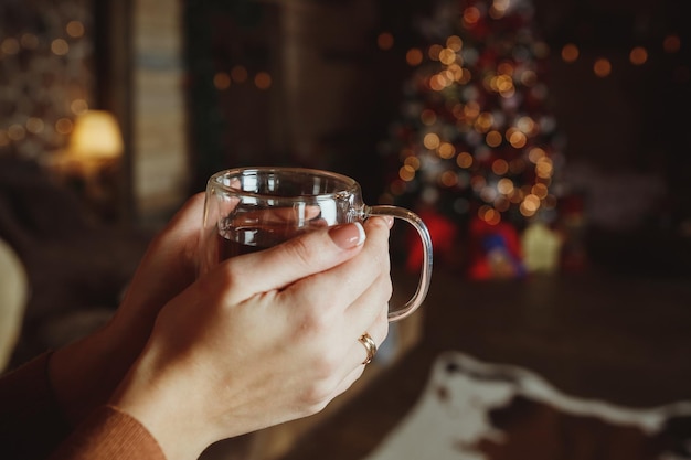 Frau, die sich im Winter mit einer Tasse Gewürztee vor einem Kamin entspanntHände in einem warmen Pullover halten eine Tasse Tee in der Nähe Ich versuche, meine Hände mit einem heißen Getränk zu wärmen