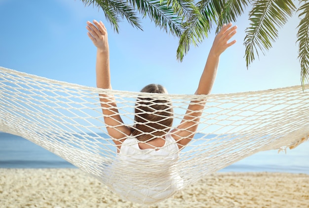 Frau, die sich in der Hängematte unter grünen Palmblättern am sonnenbeschienenen Strand entspannt