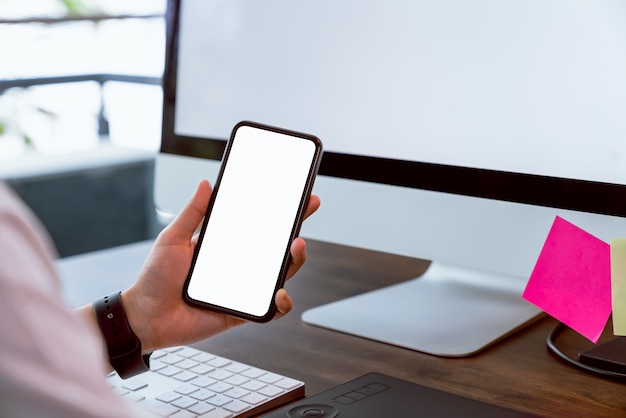 Frau, die Smartphone-Modell des leeren Bildschirms und des Computers auf dem Tischbüro hält. Nehmen Sie Ihren Bildschirm, um Werbung zu schalten.