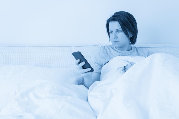 Frau, die Smartphone verwendet, wie sie im Bett sitzt, das mit Daunendecke bedeckt wird