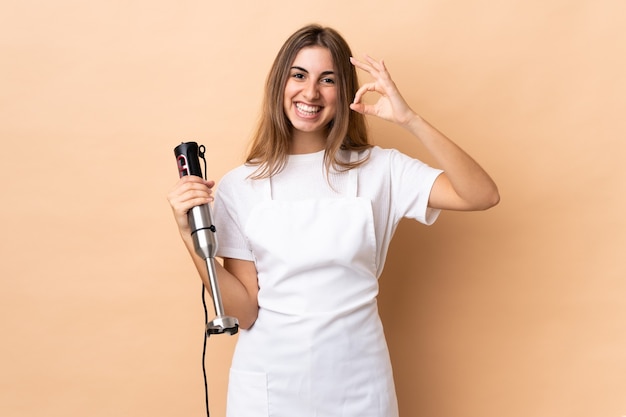 Frau, die Stabmixer über isolierter Wand verwendet, die ok Zeichen mit den Fingern zeigt