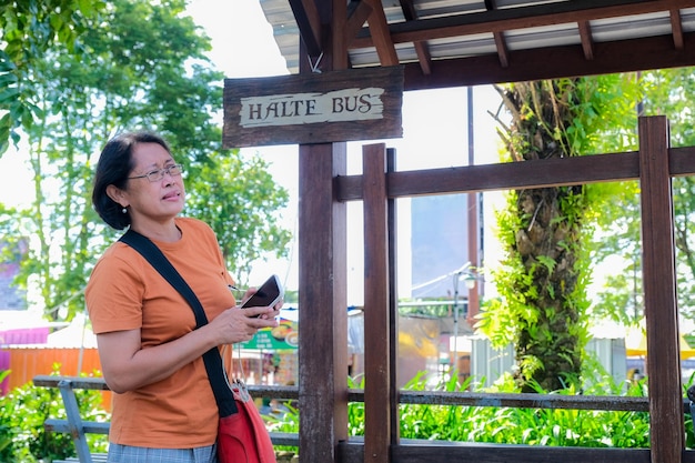 Frau, die steht und ihr Smartphone hält und an der Bushaltestelle auf ein Taxi wartet