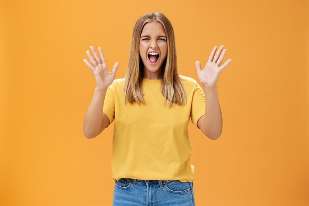 Frau, die Stress loslässt, der mit Freude und Vergnügen schreit, die mit erhobenen Armen gegen orange Hintergrund gestikulieren