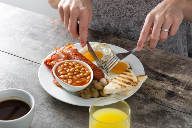 Frau, die traditionelles volles englisches Frühstück mit Spiegeleiern, Würsten, Bohnen, Pilzen, gegrillten Tomaten und Speck auf Holzoberfläche isst