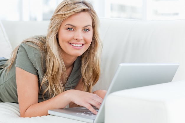 Foto frau, die über der couch liegt und lächelt, wie sie vorwärts schaut, ihren laptop verwendend