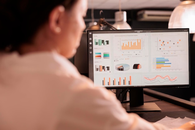 Frau, die Umsatzstatistiken für Marketingprojekte von Unternehmen auf dem Computerbildschirm analysiert und nachts im Coworking Space an Unternehmensforschung arbeitet. Unternehmensmitarbeiter überprüfen Analysen