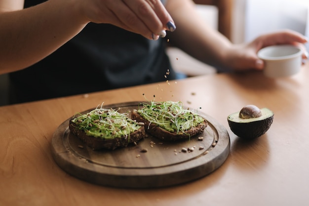 Frau, die veganes Sandwich mit geröstetem Roggenbrot mit Guacamole-Sesam und Sonnenblumenkernen macht. Frau mit Sesam auf Sandwich bestreuen