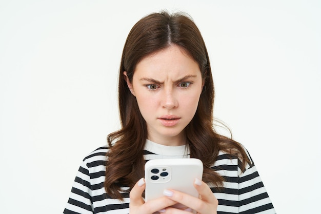 Frau, die verwirrt und schockiert vor dem Smartphone-Bildschirm sieht, die die Stirn runzelt und beunruhigende Nachrichten liest