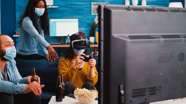 Frau, die Videospiele mit einem Virtual-Reality-Headset spielt, das während der sozialen Pandemie eine Gesichtsmaske trägt, um Krankheiten mit Covid zu verhindern, die Zeit mit Freunden verbringen, die Bier trinken