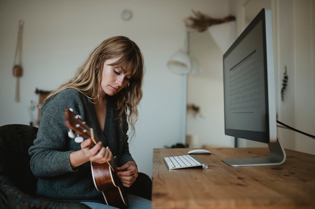 Frau, die während der Quarantäne einen Computer benutzt, während sie Ukulele spielt