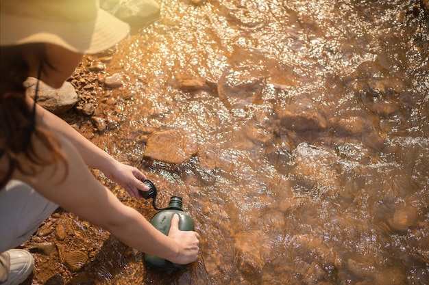 Frau, die während des Trekkings in den Bergen reines Wasser aus dem Bergbach in die Flasche nimmt Wanderer, der auf Felsen kauert und die Flasche mit kaltem Bergwasser füllt