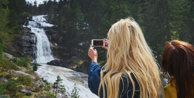 Frau, die während einer Wanderung in der Natur mit einem Freund einen Wasserfall mit ihrem Handy fotografiert