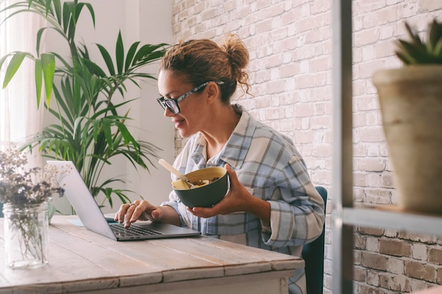 Frau, die zu Hause am Bürotisch arbeitet und gesunde Lebensmittel in einer Schüssel isst Unabhängiger weiblicher Lebensstil Alternativer Beruf mit Laptop-Computer und Online-Möglichkeit Modernes Arbeitnehmergeschäft