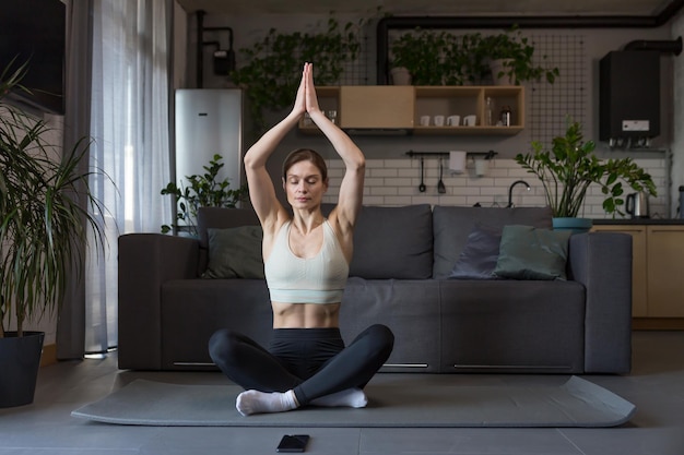 Frau, die zu Hause Yoga macht, sitzt in Lotusposition auf dem Boden und nutzt das Telefon für Online-Fitness- und Yoga-Kurse