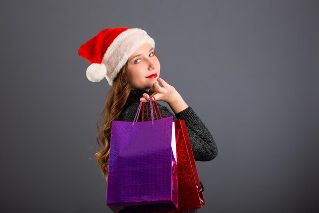 Frau einkaufen für Weihnachtsgeschenke Junge kaukasische Mädchen suchen lächelnd mit Einkaufstüten und Weihnachtsmütze Kopieren Sie Platz auf der Seite isoliert auf grauem Hintergrund