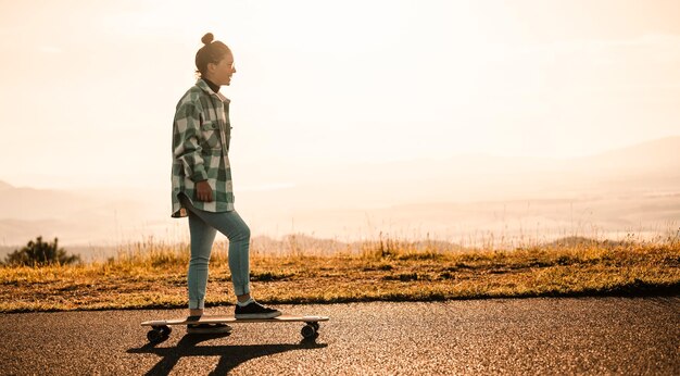 Frau fährt bei Sonnenuntergang auf gerader Straße auf Longboard Skater in Freizeitkleidung Training an Bord bei Sonnenuntergang mit orangefarbenem Licht Mädchen halten Longboard in den Händen