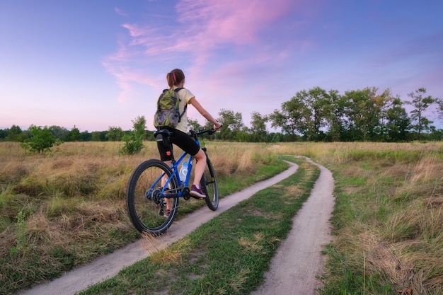 Frau fährt im Sommer bei Sonnenuntergang ein Mountainbike in der Cross Country Road. Bunte Landschaft mit sportlichem Mädchen mit Rucksackfahrrad, Feld, Schotterstraße, grünem Gras, lila Himmel. Sport und Reisen