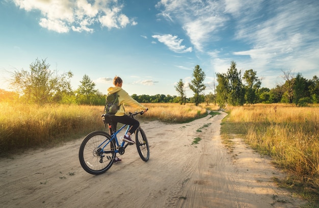 Frau fährt im Sommer bei Sonnenuntergang ein Mountainbike in der Cross Country Road. Bunte Landschaft mit sportlichem Mädchen mit Rucksackfahrrad, Feld, Schotterweg, grünes Gras, blauer Himmel. Sport und Reisen