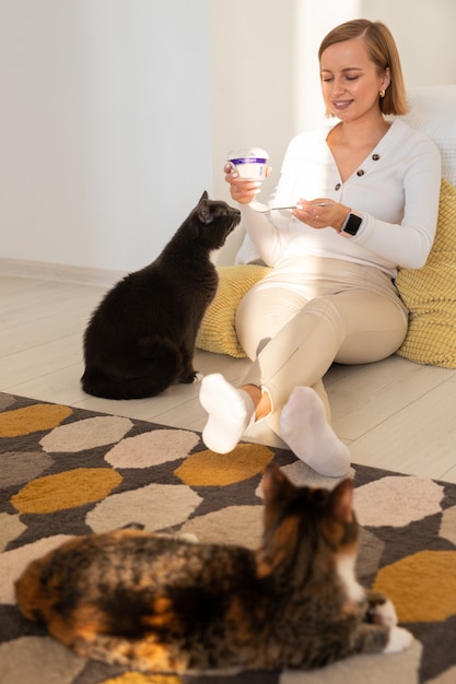 Frau füttert eine Katze mit Joghurt von einem Löffel, der auf dem Boden sitzt. Leben zu Hause mit ihren Haustieren