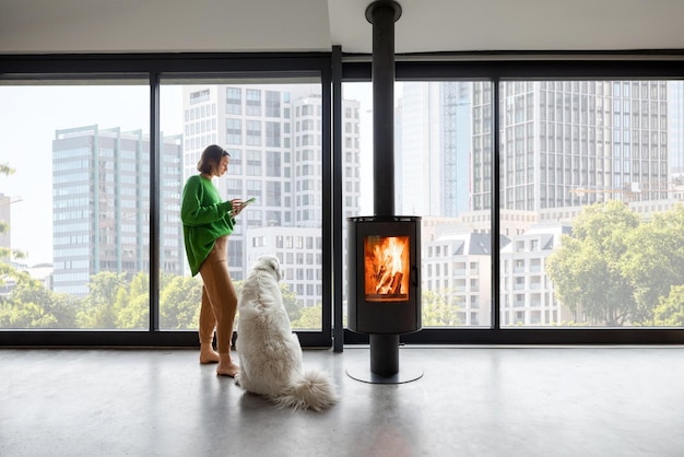 Frau genießt eine tolle Aussicht auf das Stadtbild, während sie mit Hund in der Nähe eines Kamins in einer modernen Wohnung steht