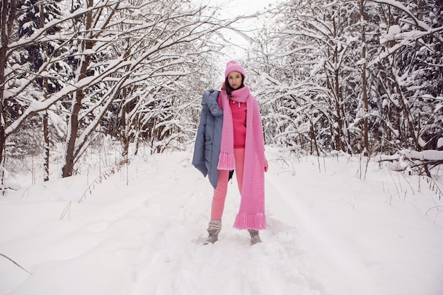 Frau genießt in rosa Kleidern eine Jacke, einen gestrickten Schal und eine Mütze, die im Winter in einem verschneiten Wald steht