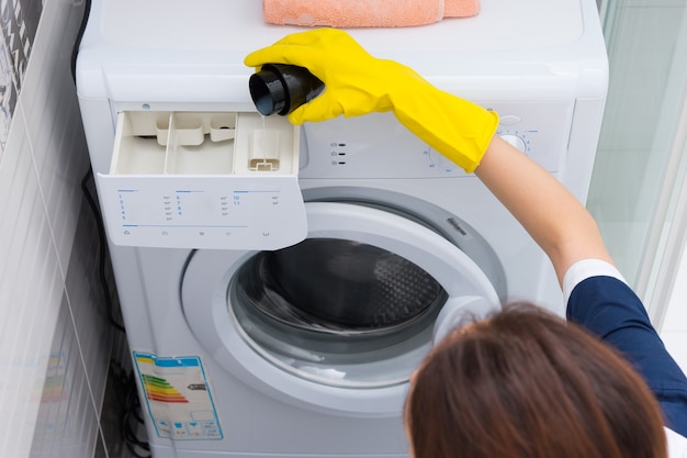 Frau gießt Weichspüler in eine Waschmaschine mit behandschuhten Händen, während sie die Hausarbeit macht, Nahaufnahme aus dem hohen Winkel