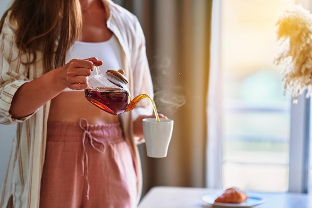 Frau gießt zu Hause heißen aromatischen schwarzen Tee aus der Teekanne aus Glas in eine Tasse