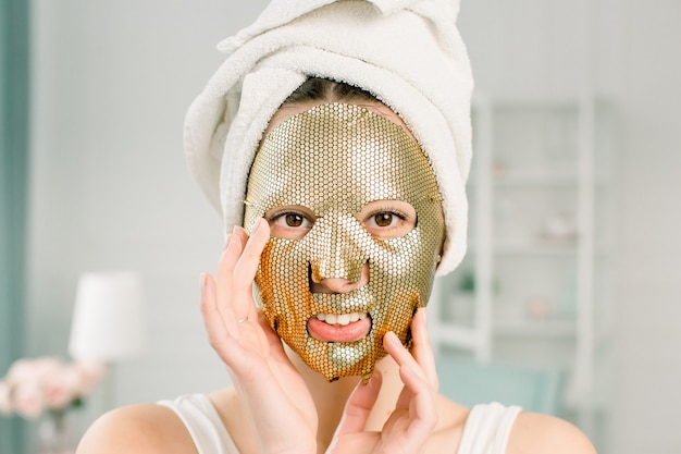 Frau Gold Sheet Mask. Schöne junge Frau im weißen Handtuch auf dem Kopf mit goldener Hautkosmetik, Kamera betrachtend. Schönheitspflege und -behandlung