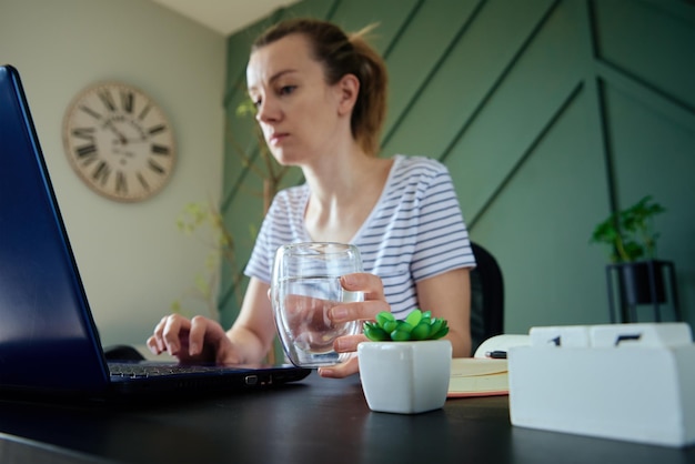 Frau hält ein Glas Wasser, während sie zu Hause am Arbeitsplatz arbeitet