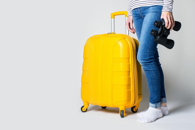 Frau hält einen gelben Plastikkoffer und ein Fernglas auf einem hellen Raum. Reisekonzept, Flugerwartung, Urlaub.