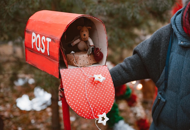 Frau hält einen roten Tupfen-Retro-Postfach mit Weihnachtsgeschenkspielzeug sowie einen Glückwunschbrief vom Weihnachtsmann auf einem Hintergrund von Tannen.