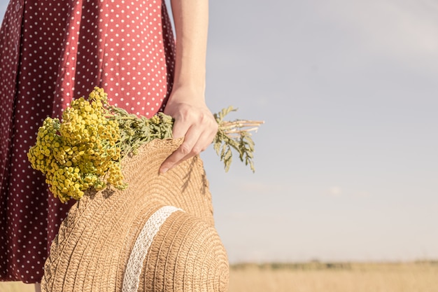 Frau hält Feldblumenstrauß und einen Hut. Ländliche Szene: Großaufnahme der Frau im Tupfenkleid mit Landwirthut und Blumenstrauß in ihrer Hand