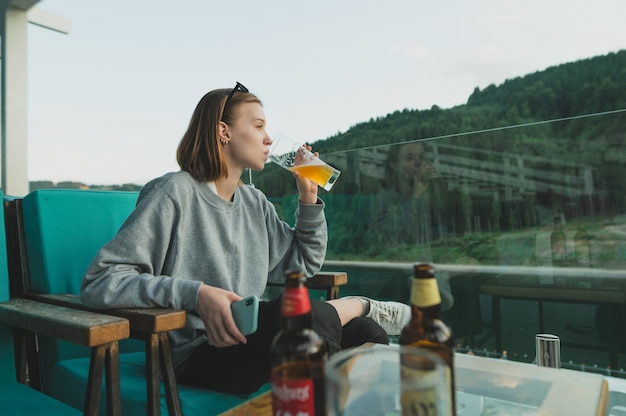 Frau hält Glas kaltes Bier im Sommer auf Terrasse an einem Restauranttisch nahe Bergwald