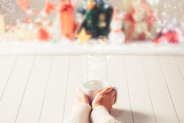 Frau hält in ihren Händen rote Kaffeetasse auf unscharfem Weihnachtshintergrund. Warmtonfotografie. Kaffeethema