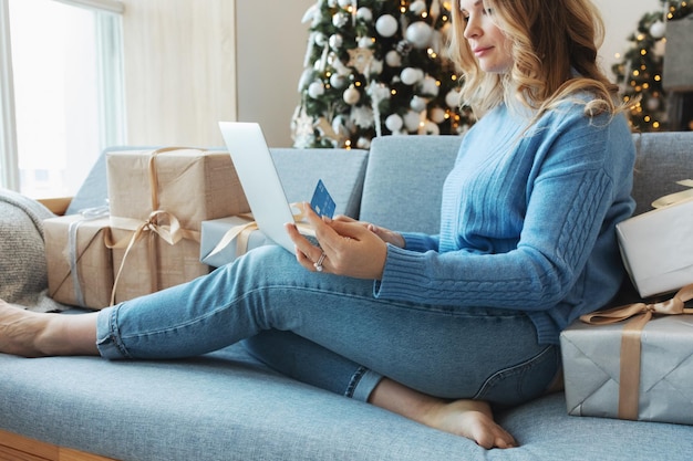 Frau hält Kreditkarte und Laptop Online-Shopping Saisonverkauf kaufen Weihnachtsgeschenke für Familie Weihnachten Hintergrund Baum Geschenkboxen Lichter Blaue Farben