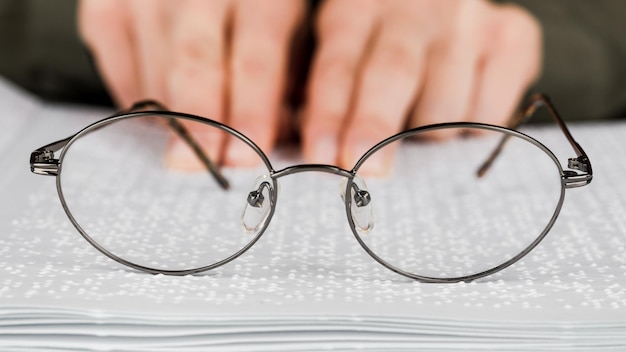 Frau hält schwarz umrandete Brille vor dem Hintergrund eines offenen Lehrbuchs, das Bücher in Blindenschrift liest