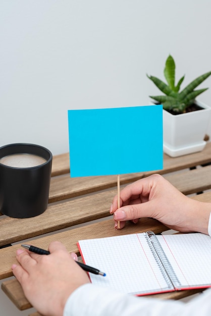 Frau hält Stift und wichtige Nachricht auf Stick auf Tisch mit Notizbuchpflanze und Kaffee sitzend