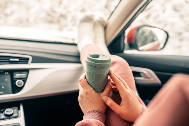 Frau hält Tasse Kaffee im Auto. Reiselebensstil. Beine am Armaturenbrett.