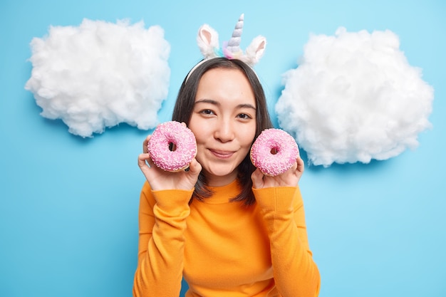 Frau hält zwei glasierte Donuts in der Nähe des Gesichts lächelt sanft in orangefarbenen Pullover gekleidet genießt es leckeres süßes Dessert isoliert auf Blau zu essen