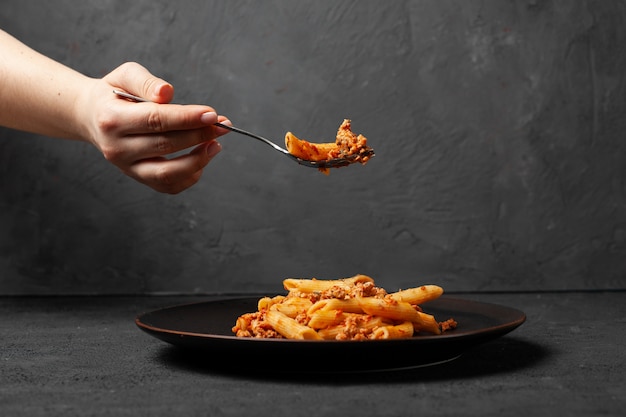 Frau halten in der Gabel frisch italienische Penne Pasta Bolognese auf dunklem Teller und dunklem Betonhintergrund.