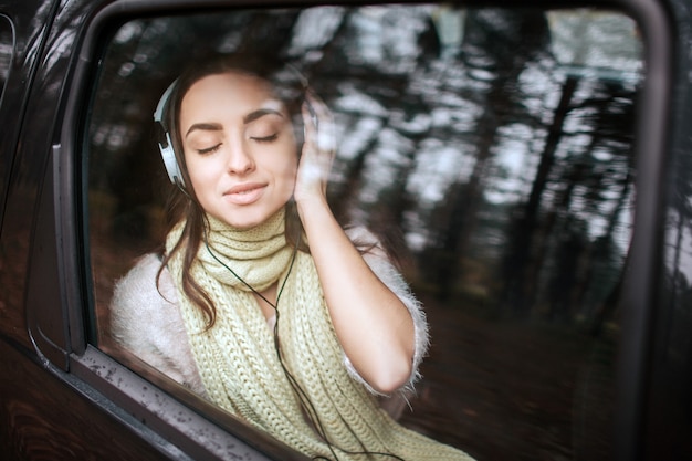 Frau im Auto, Herbstkonzept. Lächelndes hübsches Mädchen, das Musik auf Ihrem Smartphone mit Kopfhörern hört, die sich im Auto bewegen.