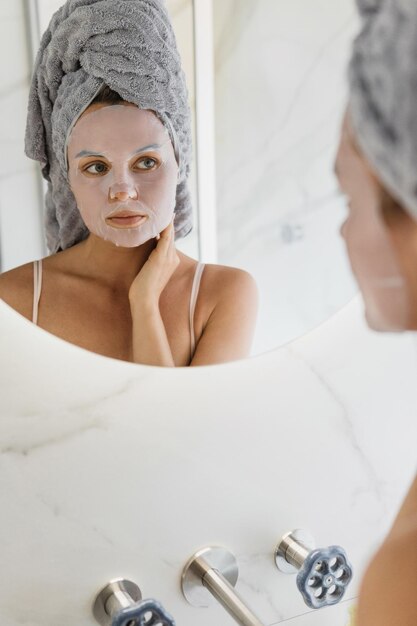 Frau im Badezimmer mit aufgesetzter Tuchmaske im Gesicht und blickt in den Spiegel