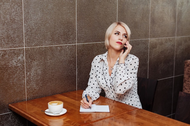 Frau im Café, die am Telefon spricht und in Notizbuch schreibt