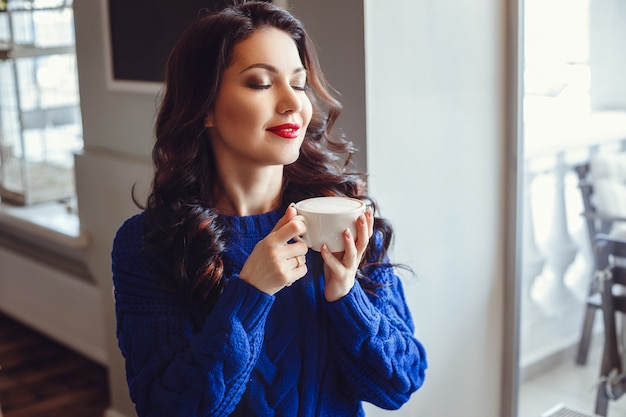 Frau im Café sitzt an einem Tisch, trinkt Kaffee und schaut zum Fenster. Frau mit rotem Lippenstift und in einem blauen Pullover wartet auf ein Treffen, telefoniert und lächelt viel.