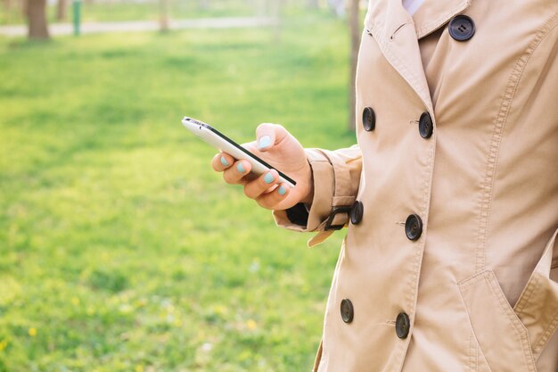 Frau im eleganten beige Mantel benutzt einen Handy im Park