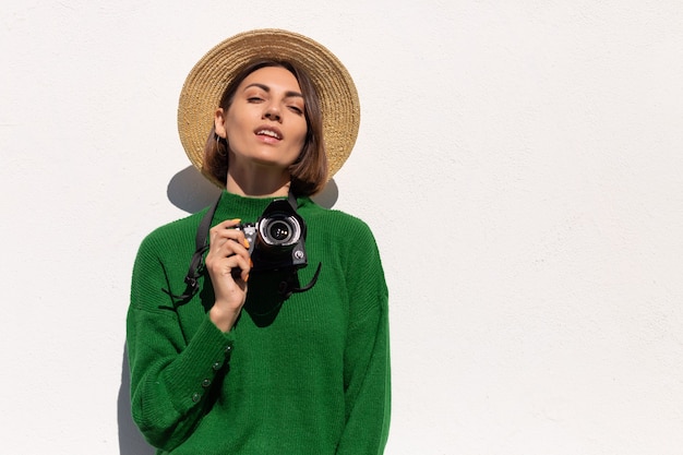 Frau im grünen lässigen Pullover und Hut im Freien auf weißer Wand glücklich positiver Tourist mit professioneller Kamera