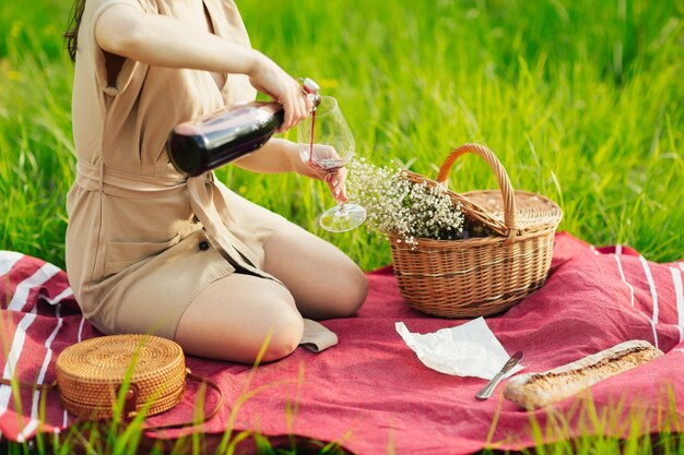 Frau im Kleid, die Wein gießt, sitzt auf einem grünen Gras draußen im Park